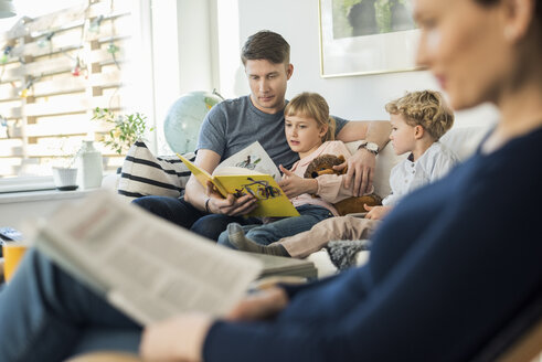 Vater liest Kindern ein Märchenbuch vor, während die Mutter im Wohnzimmer sitzt - MASF03178