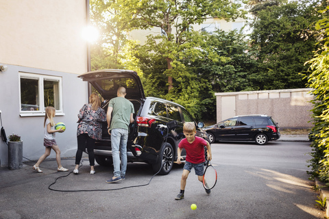 Kinder spielen mit Bällen, während die Eltern den Kofferraum ihres Autos im Hinterhof beladen, lizenzfreies Stockfoto