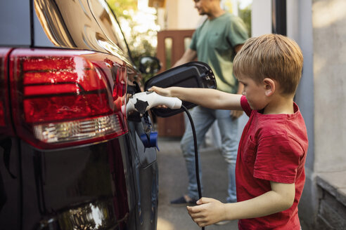 Boy charging electric car against father standing by house - MASF03244