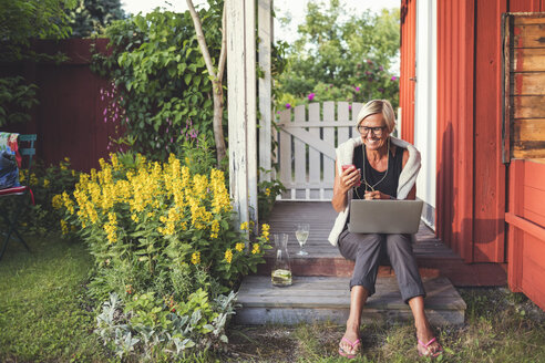 Fröhliche Frau, die ein Mobiltelefon und einen Laptop benutzt, während sie an einem sonnigen Tag im Hinterhof sitzt - MASF03247