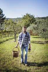 Porträt eines Mannes, der einen Korb und eine Weinflasche hält, während er auf einem Feld im Weinberg steht - CAVF36741