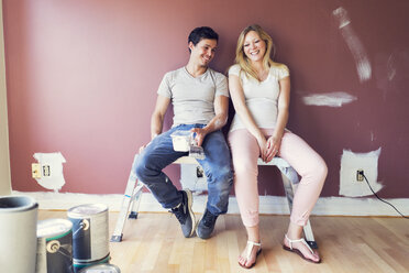 Happy couple sitting on stool against wall at home - CAVF37023