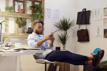 Kreativer Geschäftsmann in voller Länge, der sein Smartphone benutzt, während er im Büro sitzt und die Füße hochlegt - CAVF37076