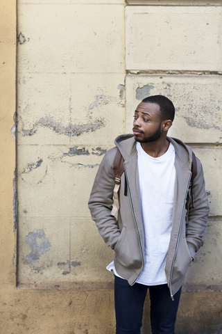 Thoughtful young man looking away while standing with hands in pockets against wall stock photo