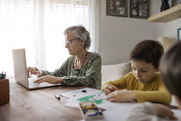 Senior woman using laptop while grandsons making drawings at home - CAVF37132