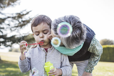 Senior woman kissing grandson playing with blowing bubbles in park - CAVF37158