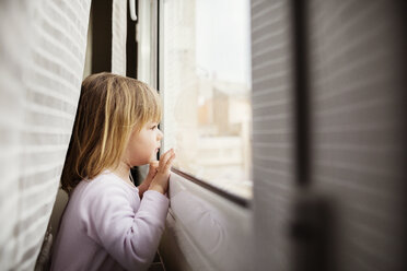 Seitenansicht eines neugierigen Mädchens, das durch ein Fenster zu Hause schaut - CAVF37172