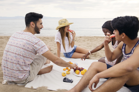 Glückliche Freunde, die am Strand sitzend Früchte essen, lizenzfreies Stockfoto