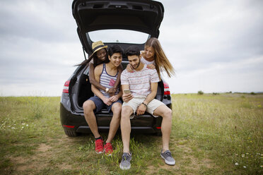 Happy friends using smart phone while sitting in car trunk on field against sky - CAVF37210