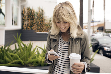 Happy young woman using smart phone while holding disposable glass by city street - CAVF37252