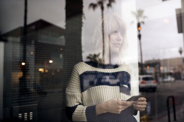 Nachdenkliche junge Frau hält Smartphone durch Glaswand gesehen - CAVF37259