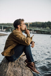 Side view of man smoking while sitting on log by river against clear sky - CAVF37268