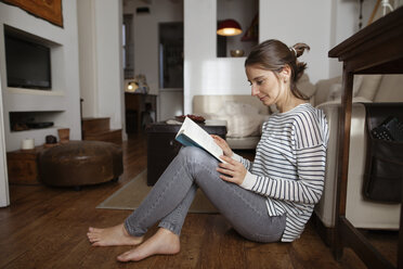 Frau liest ein Buch, während sie zu Hause auf dem Hartholzboden sitzt - CAVF37289