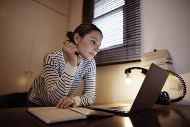 Frau arbeitet an Laptop bei Schreibtischlampe am Tisch - CAVF37298