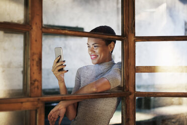 Smiling businesswoman using phone while leaning on window - CAVF37330