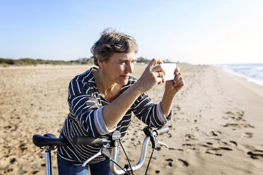 Frau beim Fotografieren mit Fahrrad am Strand stehend - CAVF37336
