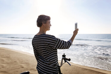 Frau fotografiert mit dem Mobiltelefon, während sie mit dem Fahrrad am Strand steht - CAVF37337