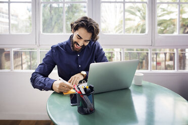 Glücklicher Geschäftsmann, der auf einem Klebezettel schreibt, während er am Tisch mit einem Laptop im Büro sitzt - CAVF37387