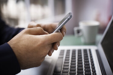 Nahaufnahme der Hände eines Geschäftsmannes, der ein Smartphone über einem Laptop in einem kreativen Büro benutzt - CAVF37389