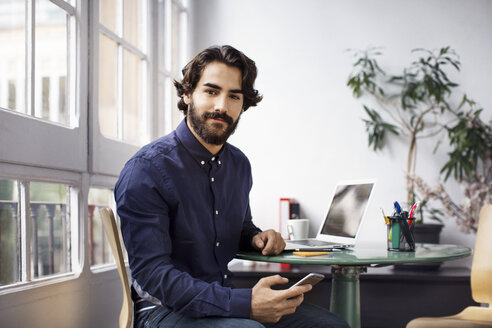 Porträt eines zuversichtlichen Geschäftsmannes, der ein Smartphone hält, während er im Büro sitzt - CAVF37390