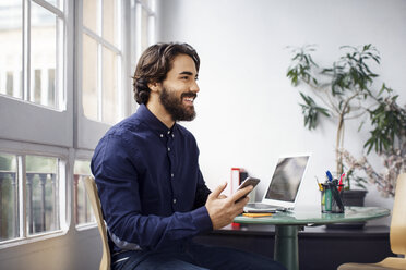 Nachdenklicher Geschäftsmann, der sein Smartphone hält und auf einem Stuhl im Büro sitzt - CAVF37391