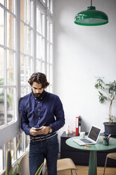 Businessman using mobile phone while leaning on window in office - CAVF37393