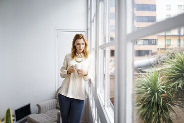 Thoughtful businesswoman leaning on window while holding coffee cup in office - CAVF37396