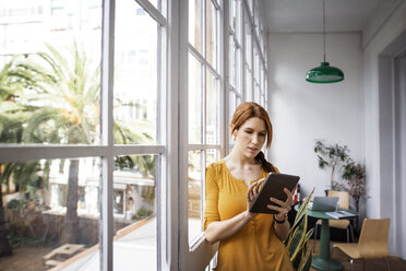 Businesswoman using tablet computer while leaning on window in office - CAVF37409