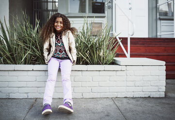 Porträt eines glücklichen Mädchens, das auf einer Stützmauer am Bürgersteig sitzt - CAVF37560