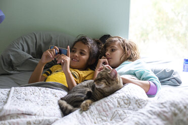 Schwestern schauen auf ihr Smartphone, während sie mit ihrer Katze auf dem Bett liegen - CAVF37599