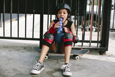 Verspieltes Mädchen trinkt Wasser, während es auf einem Skateboard gegen ein Geländer sitzt - CAVF37606