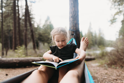 Abgeschnittene Beine der Mutter um die Tochter, die in der Hängematte auf dem Campingplatz ein Märchenbuch liest - CAVF37658