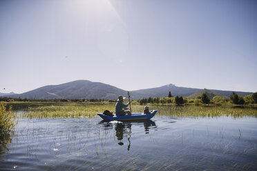 Father and daughter sitting in inflatable raft on lake against clear sky during summer - CAVF37685