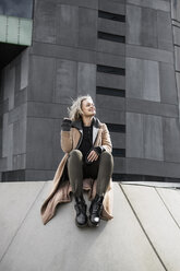 Glückliche Frau sitzt auf einer Stützmauer vor einem grauen Gebäude - CAVF37742