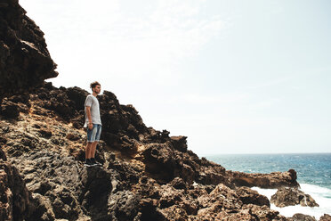 Side view of young man standing on rocks by sea against sky - CAVF37755