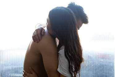 Seitenansicht eines romantischen Paares, das sich umarmt, während es zu Hause am Fenster steht - CAVF37804