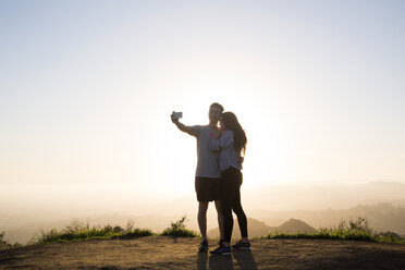 Couple taking selfie through smart phone while standing on mountain against clear sky - CAVF37809