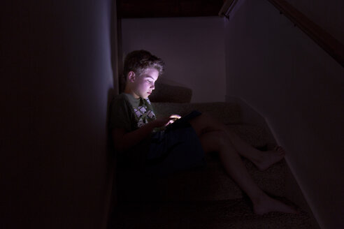 Junge spielt Spiel auf Tablet-Computer, während er zu Hause auf den Stufen sitzt - CAVF37830