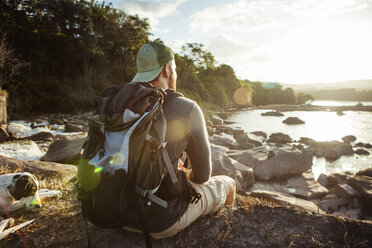 Rear view of male hiker sitting on rocky lakeshore during sunset - CAVF37975