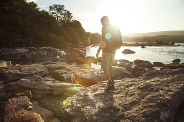 Side view of male hiker walking on rocky lakeshore during sunset - CAVF37977