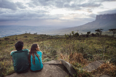 Rückansicht eines Mannes und einer Frau, die auf einem Felsen vor einem bewölkten Himmel sitzen - CAVF37998