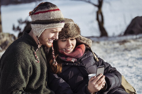 Lächelndes Paar, das ein Smartphone benutzt, während es sich auf einem verschneiten Feld entspannt - MASF03265