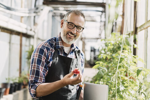 Smiling gardener with digital tablet holding tomato in greenhouse - MASF03292