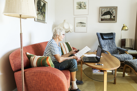 Ältere Frau analysiert ein Dokument, während sie zu Hause einen Laptop benutzt, lizenzfreies Stockfoto