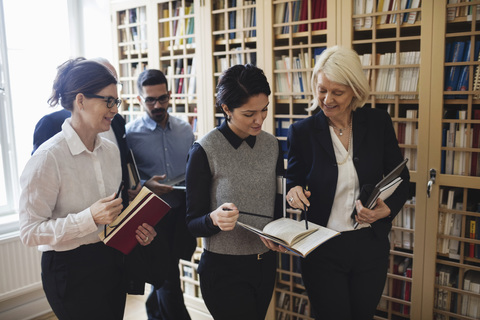 Lächelnde Anwälte diskutieren, während sie an einem Bücherregal in einer Bibliothek vorbeigehen, lizenzfreies Stockfoto