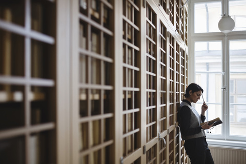 Thoughtful female lawyer researching while leaning on shelf in library stock photo