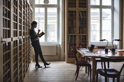 Nachdenklicher Anwalt hält ein Buch und lehnt sich an ein Regal in der Bibliothek - MASF03336