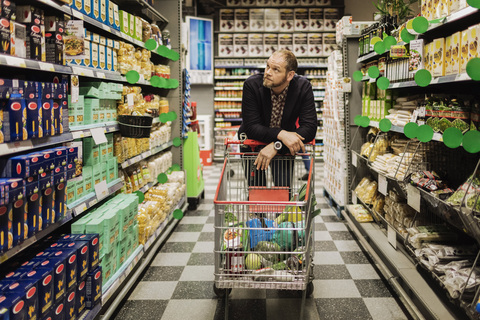 Nachdenklicher männlicher Kunde, der sich im Supermarkt auf einen Einkaufswagen stützt, lizenzfreies Stockfoto