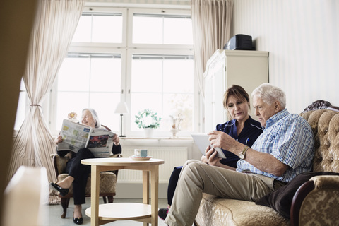 Ein älterer Mann und ein Pfleger benutzen ein digitales Tablet, während die Frau im Pflegeheim Zeitung liest, lizenzfreies Stockfoto