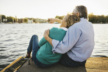 Senior man showing something to woman while sitting on pier - MASF03401